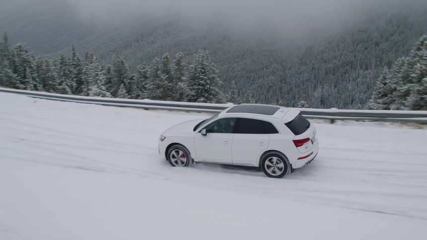 A white car driving on a snowcovered street next to a cliff with many trees