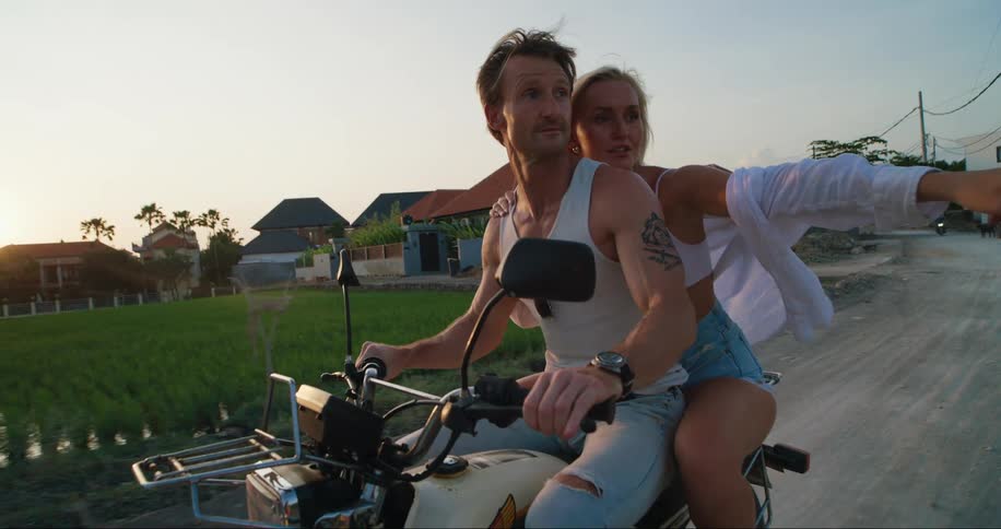 Man and woman ride motorcycle by houses and field
