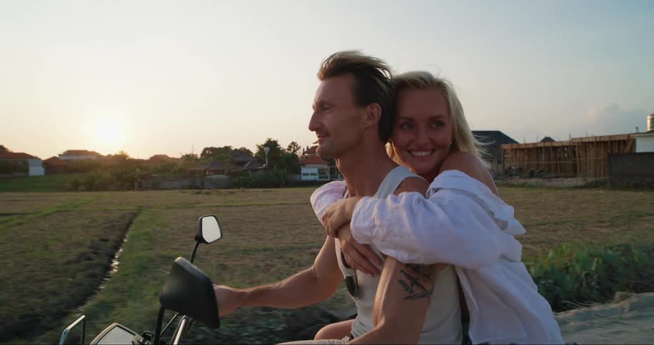 A woman smiles as she holds on to a man driving her on a motorcycle
