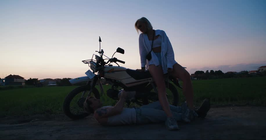Person sits on motorcycle as other person lays on ground repairing motorcycle at dusk