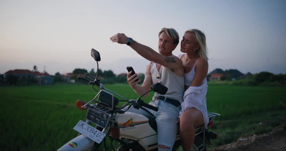 Man and woman sit on motorcycle near field looking at phone for directions