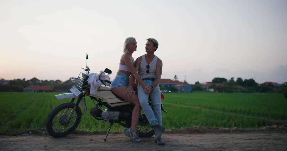 Man and woman sitting side by side affectionately on a motorcycle on the edge of an agricultural field