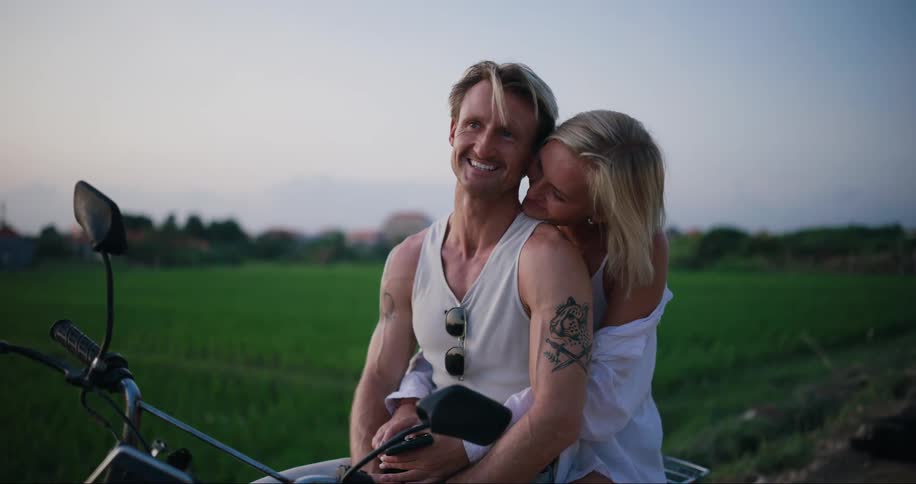 A woman kisses a man while they sit on a motorcycle by a field