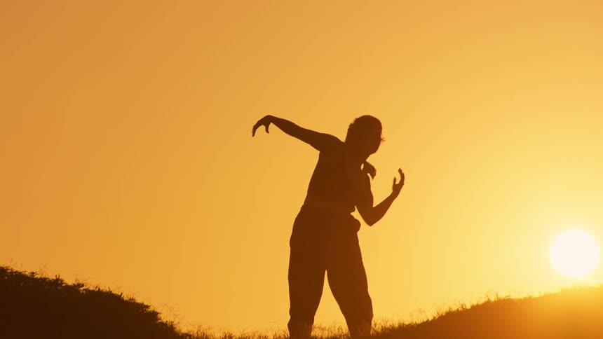 Silhouette of a woman on a mountain peak doing tai chi movements against an orange sun in the background