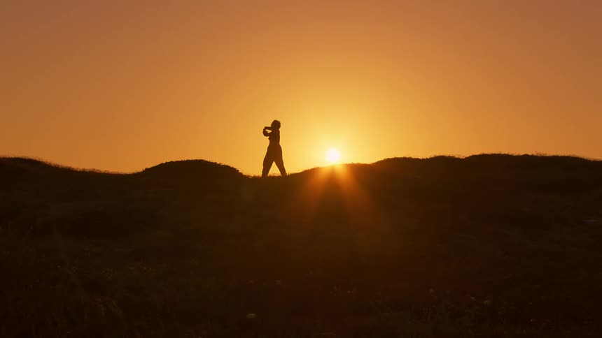 Silhouette of a person on a hill moving their arms and legs and in the background a setting sun