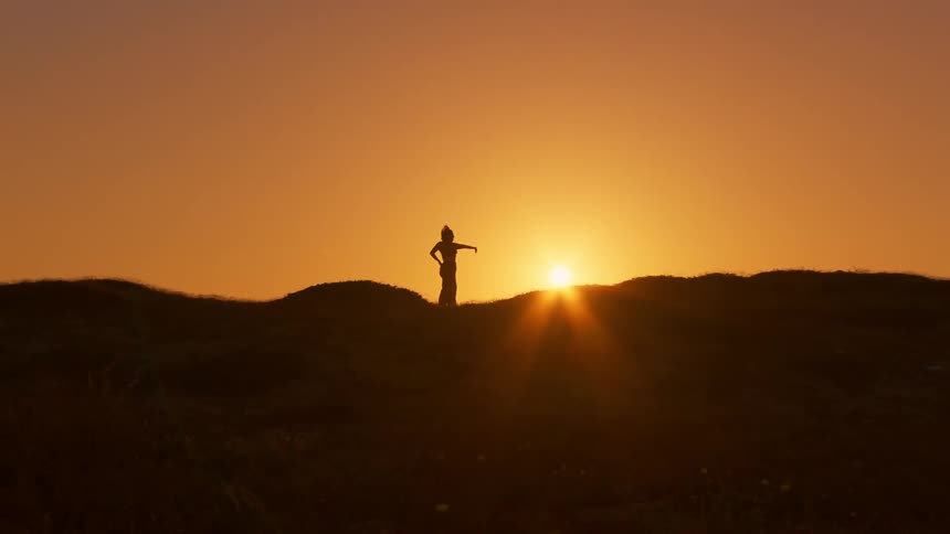 Silhouette of a person on the peak of a hill making body movements against the golden sun on the horizon