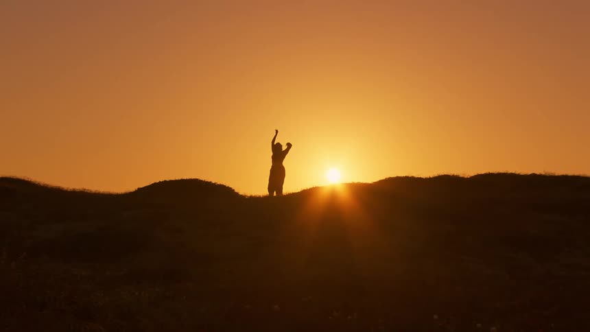 Silhouette of a person against an orange sun and on a mountain peak dancing
