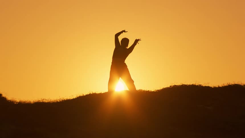 Silhouette of a person at the peak of a hill moving their arms against a golden sun
