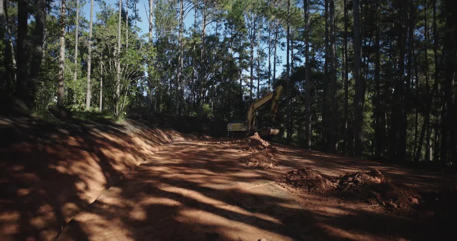 A yellow excavator moves dirt in a road through a dense forest
