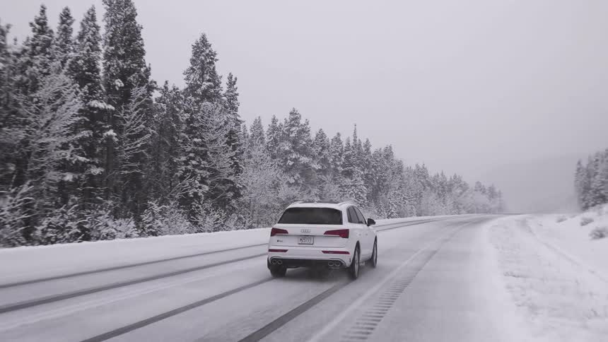 A white car drives down a snow covered street by a forest