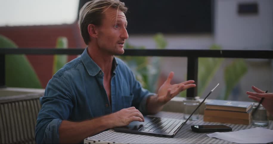 A man and a woman sit at a table on a balcony while having a discussion