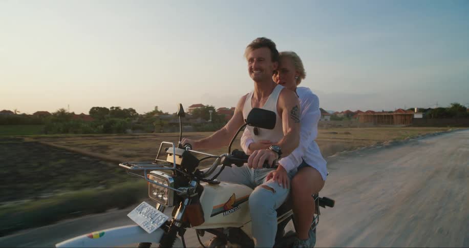 A couple rides a motorcycle down a rural dirt road