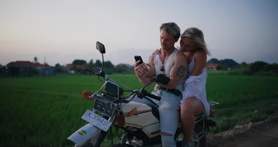 A young couple sitting on a motorcycle parked in the countryside looking at a phone at dusk