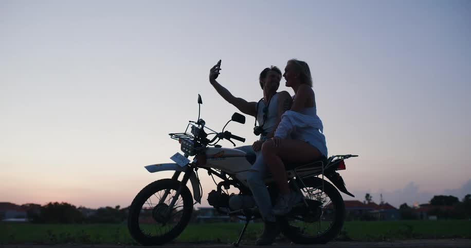 Couple sitting together on a motorcycle after dusk shares a smartphone during a video call