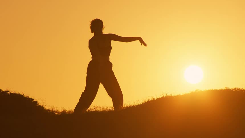 Silhouette of a woman making body movements while standing on the peak of a hill with the orange sun on the horizon