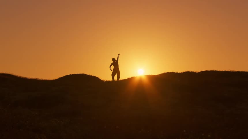 A silhouette of a woman dancing over a hill against the backdrop of a setting sun