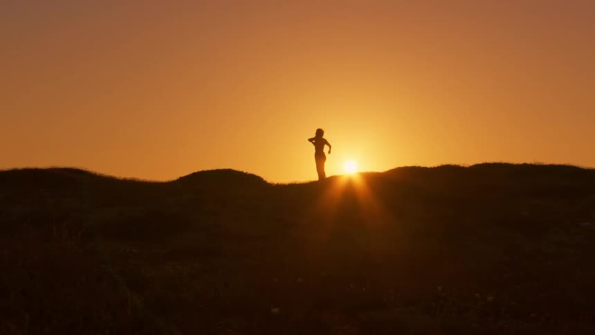 Silhouette of a person stretching and making body movements while standing on a hill against a sun on the horizon