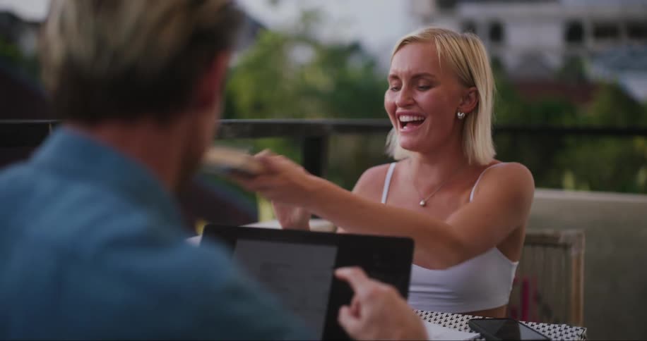 A man taps his tablet and the woman laughs then closes her book and places it on the table