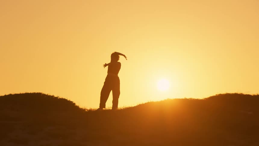 Woman dances on hill while silhouetted against the setting sun