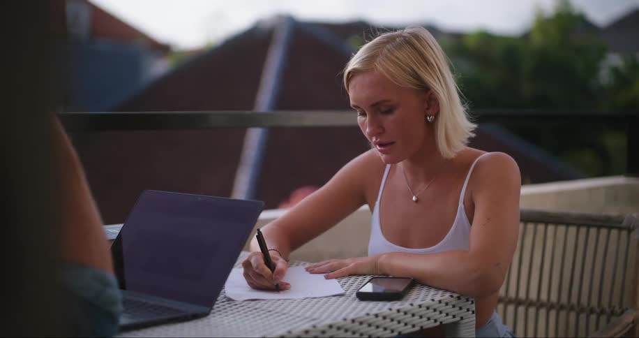 Blonde woman sitting at a table writing and talking to a person