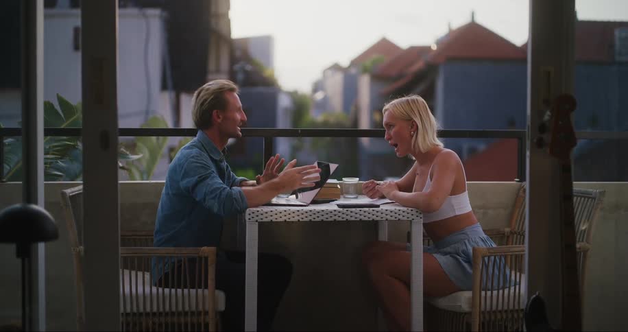 A man and a woman talk and smile while sitting together on a balcony