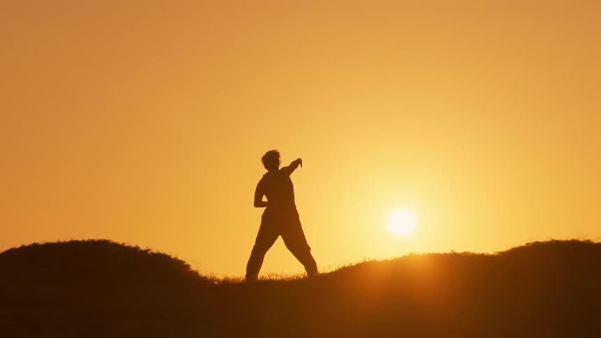 A silhouette of a woman dancing over a hill against the backdrop of a setting sun
