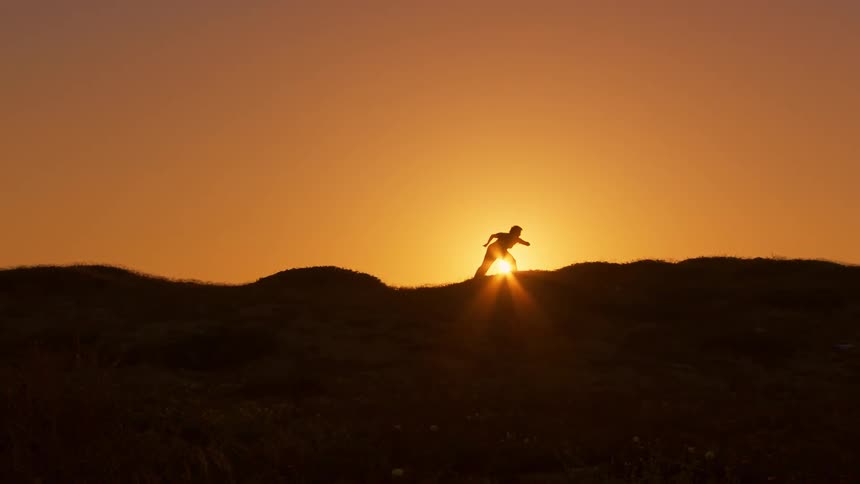 Silhouette of a person on a hill making body movements against a golden sun in the background