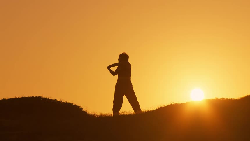 A woman is doing tai chi as the sun rises behind her