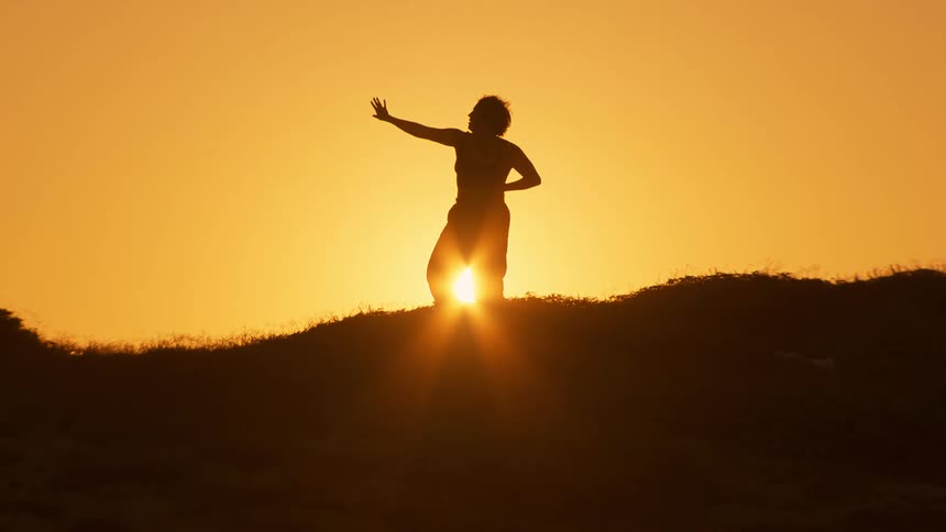 Silhouette of a person moving their body against the orange sun on a hill