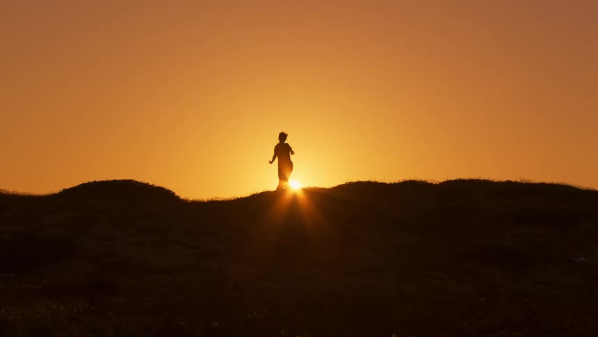 A silhouette of a person making movements with their arms and contrast with the sunlight on the horizon in orange tones in the sky