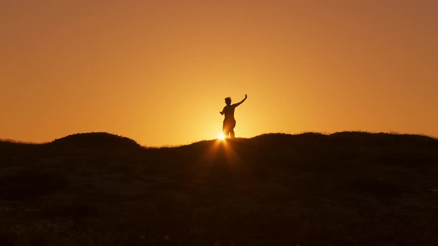 Silhouette of a woman doing taishi on a mountain peak against an orange late afternoon sun