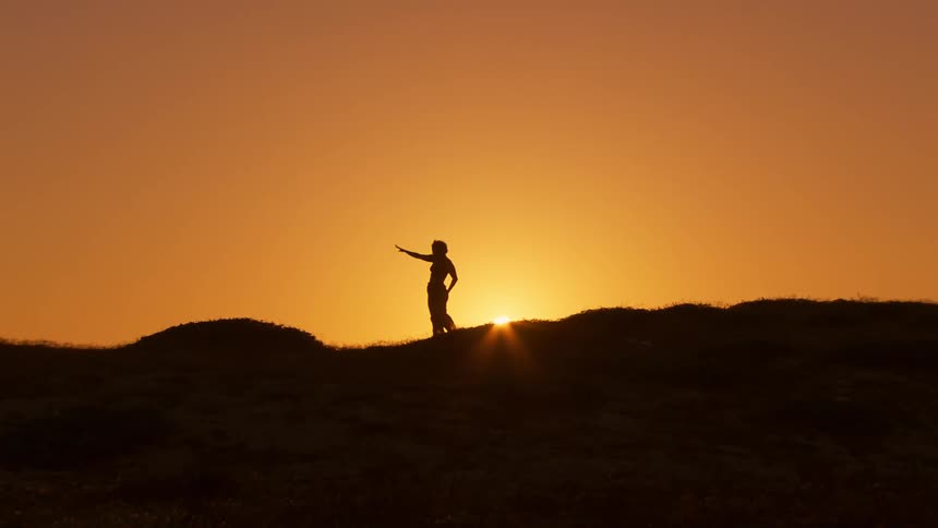Silhouette of a woman doing tai chi on a natural mountain peak against the orange sky and the sun on the horizon