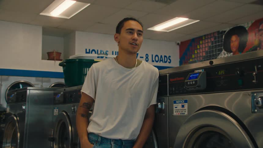 A young man in a white tshirt and jeans leans casually against a row of industrial washing machines in a laundromat smiling and appearing relaxed
