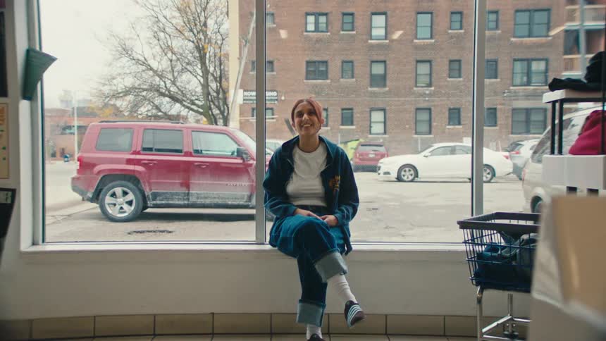 A woman smiles and gestures as she sits on the window ledge of a laundromat