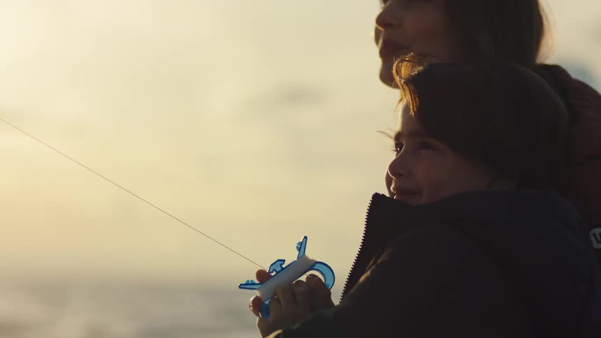 Mother and son side by side flying kites by the sea at sunset