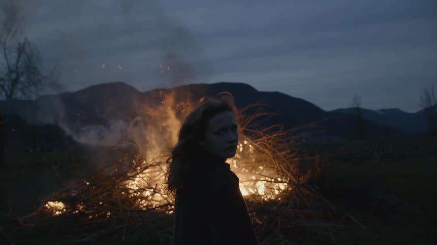 Wild haired person standing before a large outdoor brushfire on a cloudy evening