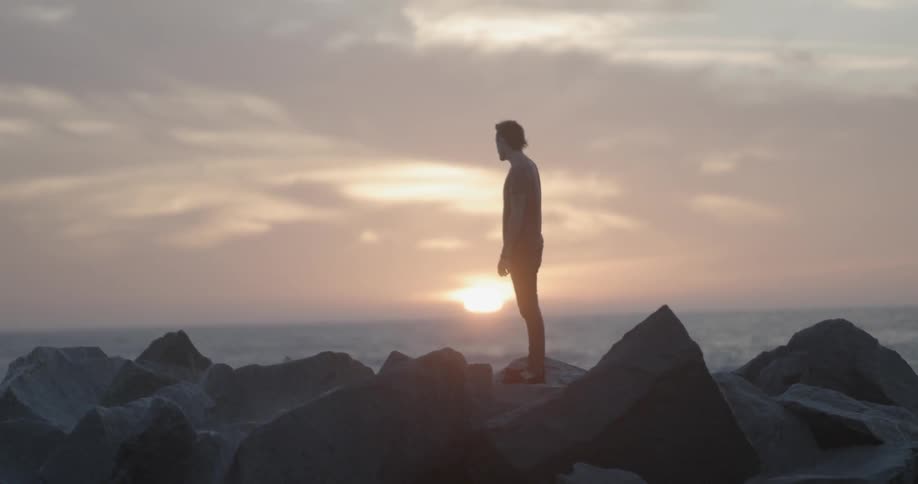 Man standing in a rocky area watching the waves come ashore with a cloudy sunset in the distance