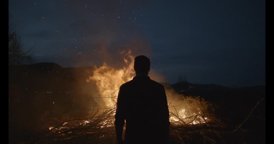 A man stands in front of a bonfire at night
