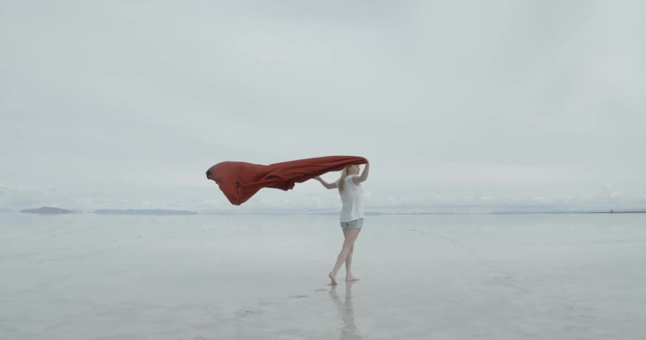 Woman standing on a beach holding a red blanket that is blowing strongly behind her