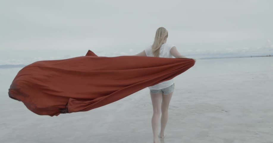 Blonde woman in a white top holding a large red cloth behind her with her arms spread wide outdoors as the wind blows strongly