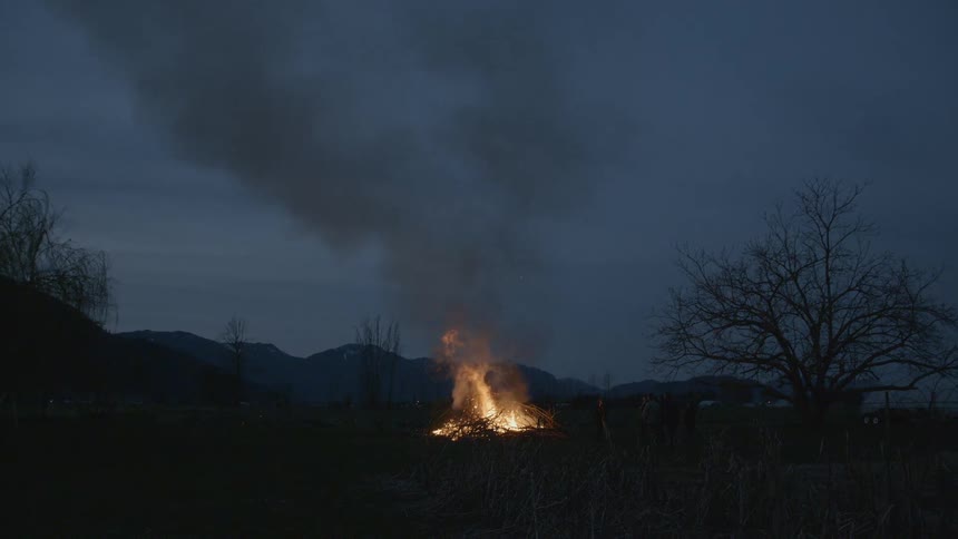 People stand looking at a bonfire burning at dusk