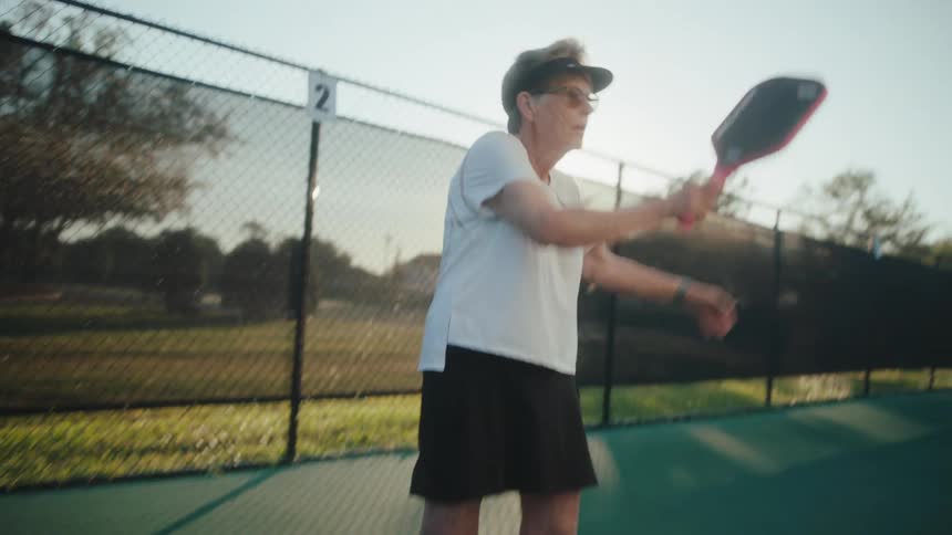 An elderly woman is hitting a pickleball with a paddle on a tennis court