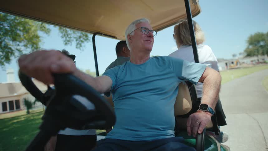Older male driver sitting in golf cart with friends talks to someone while stopped on the path