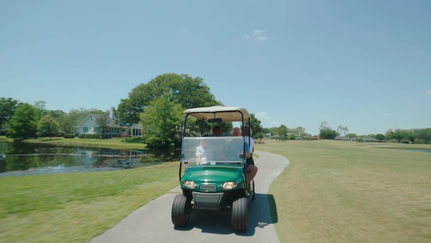 A golf cart with four passengers driving along a paved path bordered by grass and water features