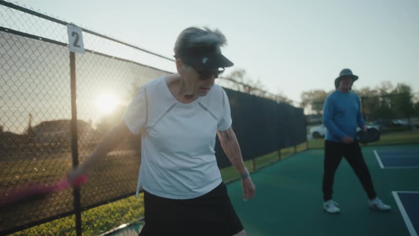 A older woman makes a pickleball serve with a yellow pickleball