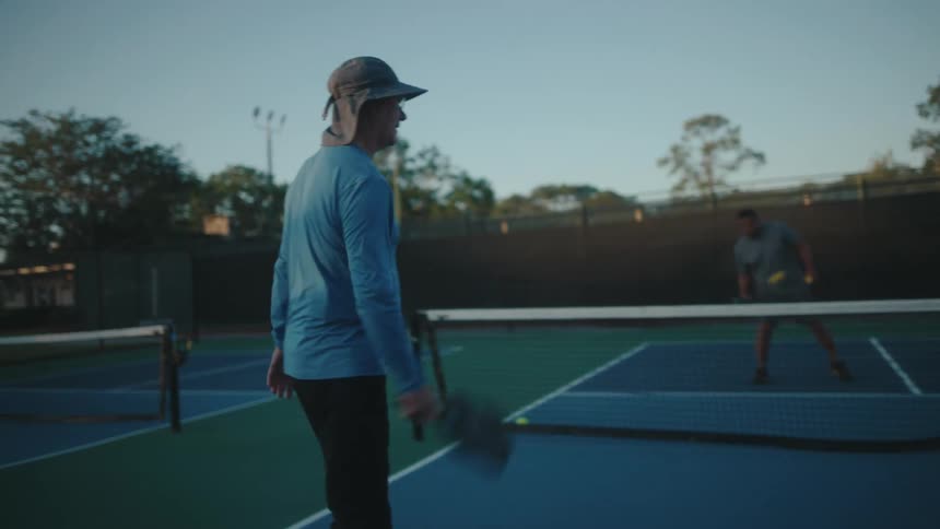 Three people hit pickleballs around on a court at dusk