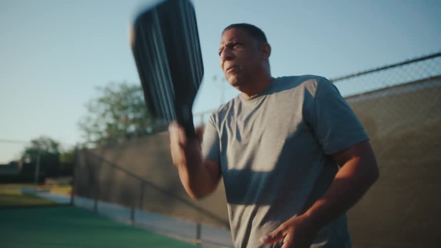 Older man playing pickle ball on a tennis court serves the ball
