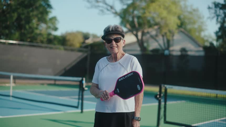 A woman holds a pickle ball paddle while smiling and standing on a playing court