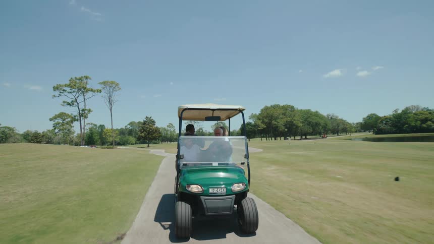 A green golf cart with four occupants driving on a path through a golf course with a clear blue sky