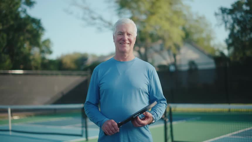 An elderly man smiling while holding a paddle and standing on a pickleball court